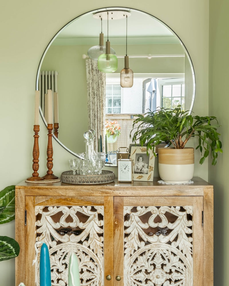 Decorative wooden cabinet with a large round mirror, plants, and candles against a light green wall.